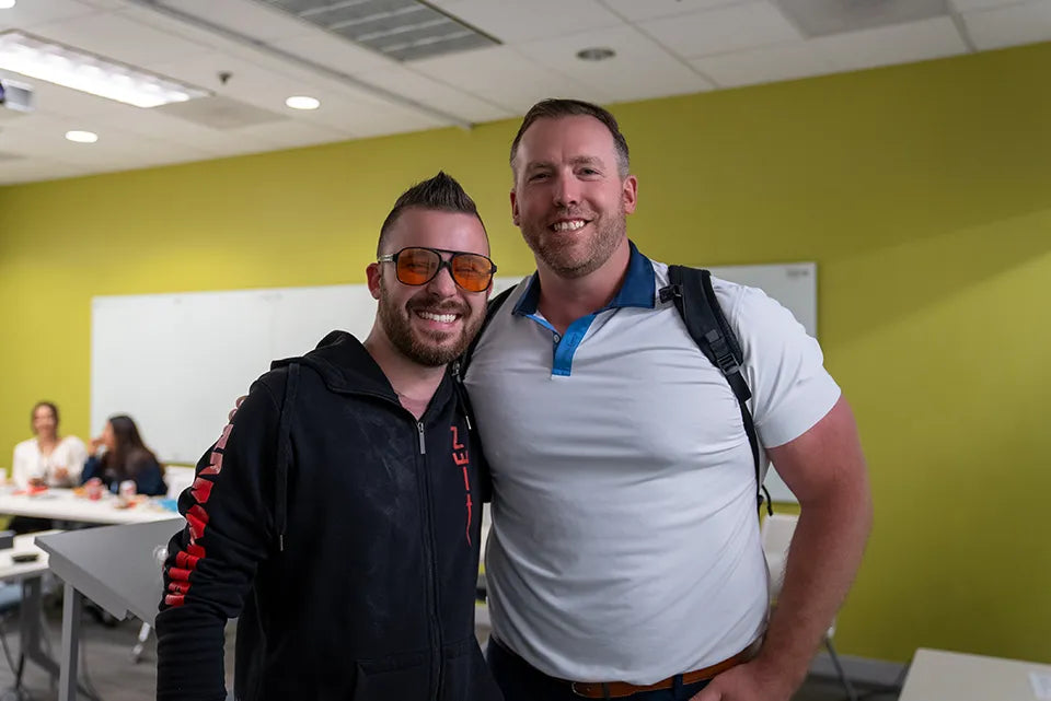 Two men posing for a photo in a room with a whiteboard and people in the background.