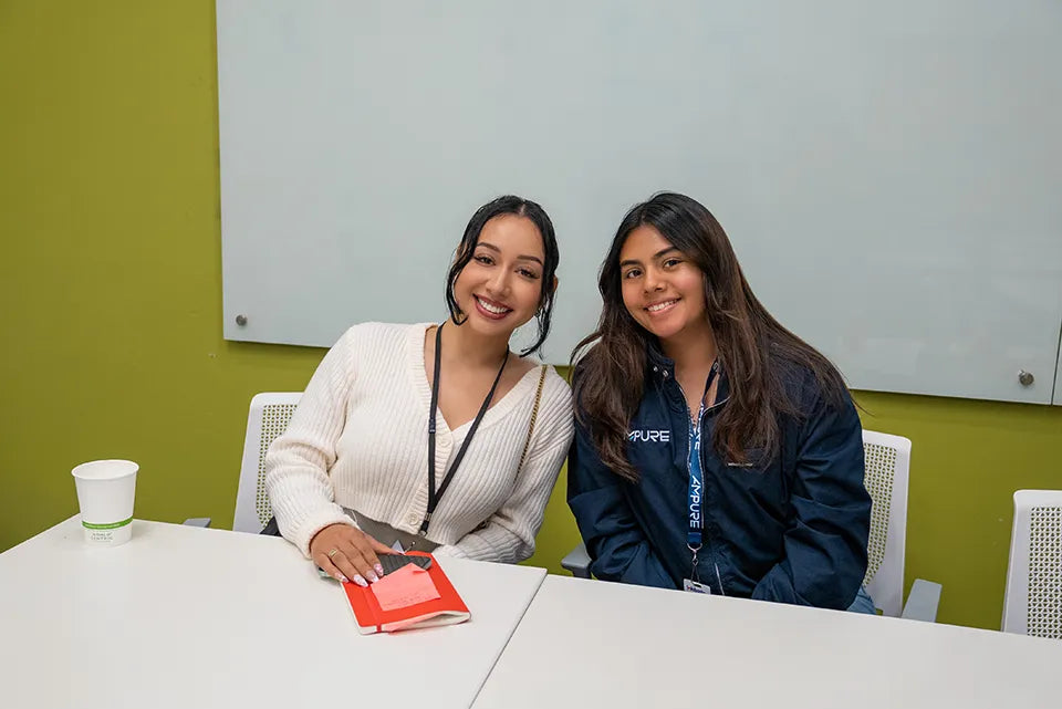 Two women sitting at a table with a green wall and whiteboard in the background