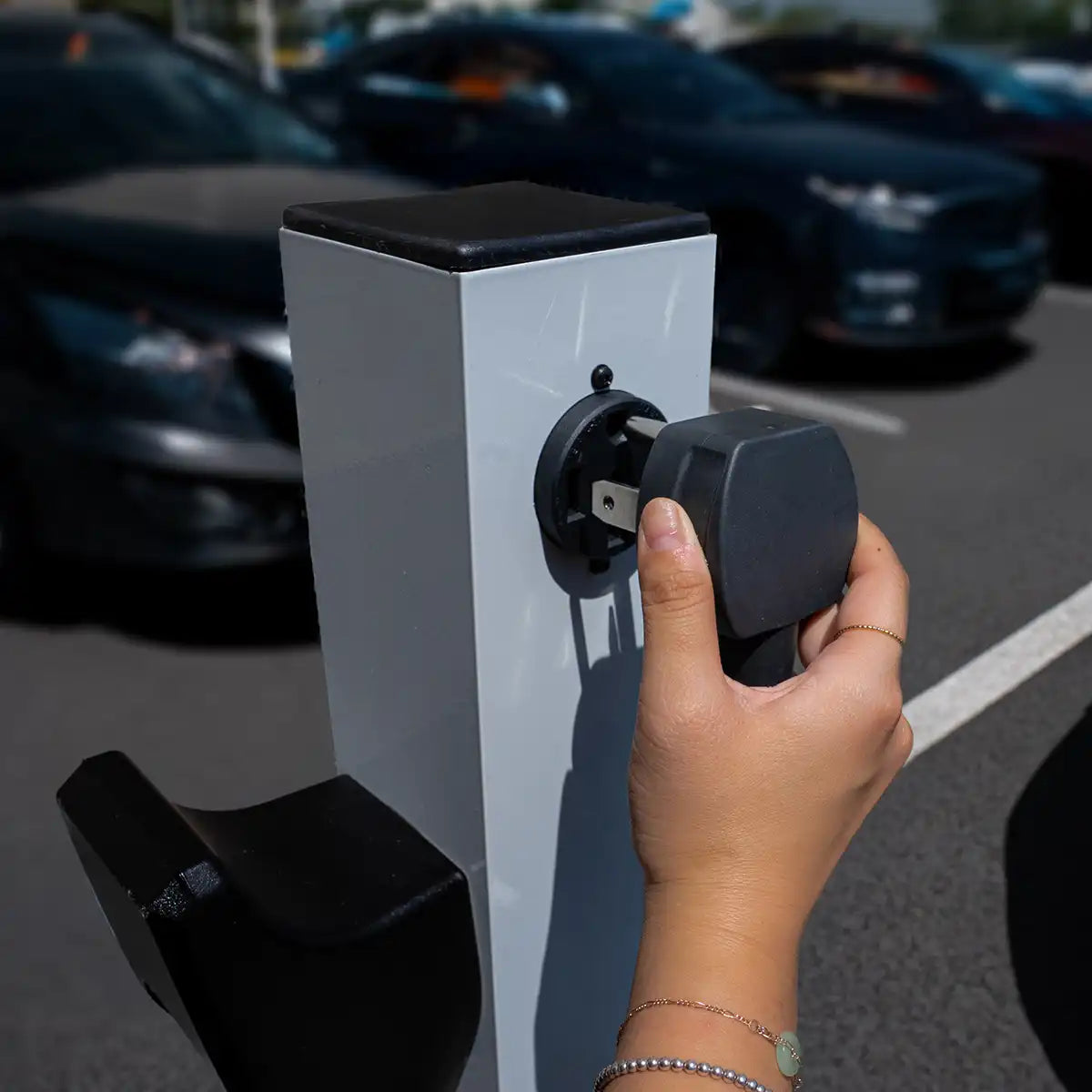 Hand plugging an electric vehicle charging cable into a charging station with cars in the background.