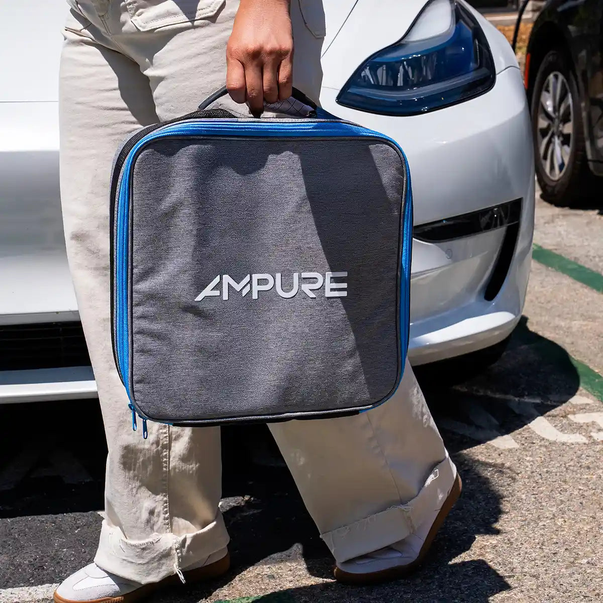 Person holding a gray and blue Ampure bag in front of a white car.